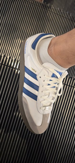 A foot wears a white adidas Samba sneaker with blue stripes and a "SAMBA" label, shown against a metallic grated floor. The shoe's clean, branded design fits the post's mention of the rare adidas × Coinbase collaboration.