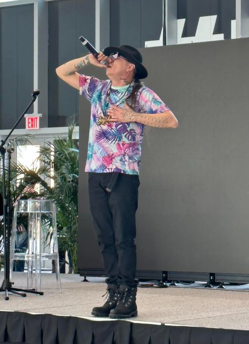 A performer with long braided hair, a black hat, sunglasses, and tattoos sings into a microphone on stage, wearing a colorful tropical-print t-shirt, black pants, and chunky black boots.