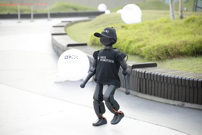 A humanoid robot wearing an "AXIS ROBOTICS" shirt and black cap walks along a curved walkway, with an OKX Wallet promotional display visible nearby. The image illustrates the Axis Robotics project behind the announced $AXIS tokenomics, including the 100 million tokens allocated for airdrop and 1 billion total token cap.