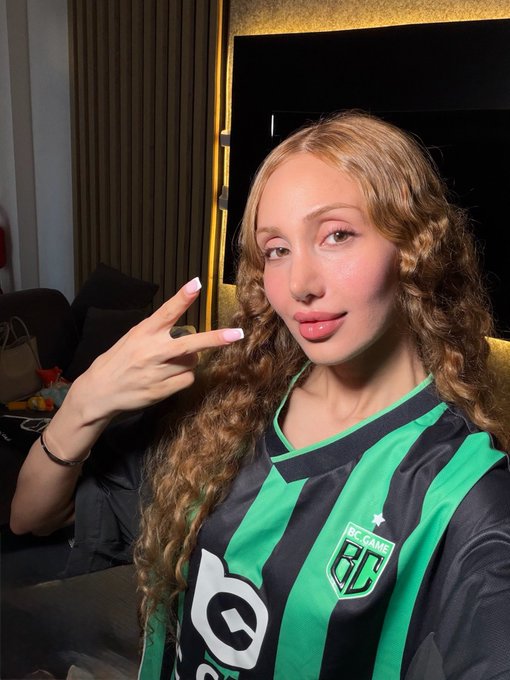 A young woman with long curly blonde-brown hair posing in a green and black striped soccer jersey featuring 'BC GAME' branding.