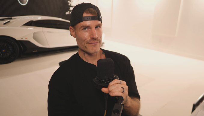 A man wearing a backwards black cap and t-shirt holds a microphone while being interviewed in front of a white Lamborghini, suggesting he is a crypto influencer promoting his latest video on Bitcoin buying strategies.