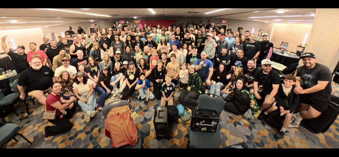 A large group photo of attendees gathered in a convention hall, posing together with various merchandise and luggage in the foreground.