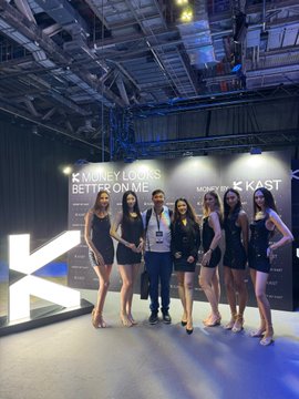 A man standing with a group of promotional models in black dresses at a booth for 'KAST' with the phrase 'Money Looks Better on Me' displayed on the wall.