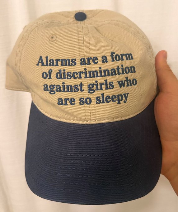 A hand holds up a two-toned beige and navy baseball cap embroidered with the humorous text 'Alarms are a form of discrimination against girls who are so sleepy'.
