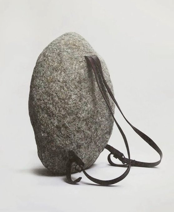 A gray speckled rock stands upright on a white surface with a brown leather strap looped around it, creating a sculptural, found-object composition.