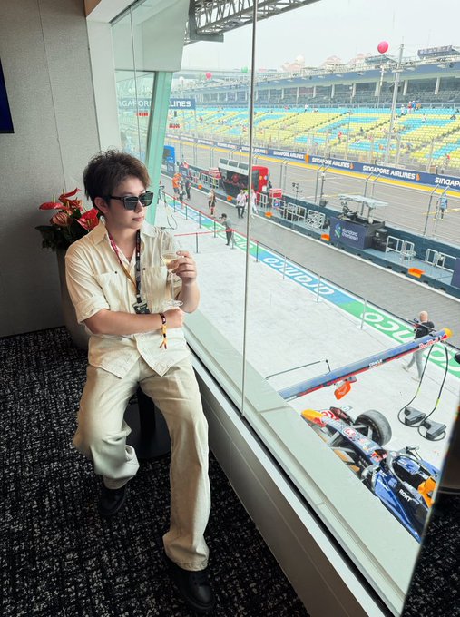 Ric, dressed in a light-colored shirt and trousers with sunglasses, stands in a suite overlooking the Singapore Grand Prix circuit, with "Singapore Airlines" and "Singapore GP" signage visible on the barriers outside the window. Below, a race car can be seen in the pit lane, connecting the image to the post's description of his experience hosting guests in the VIP paddock area.