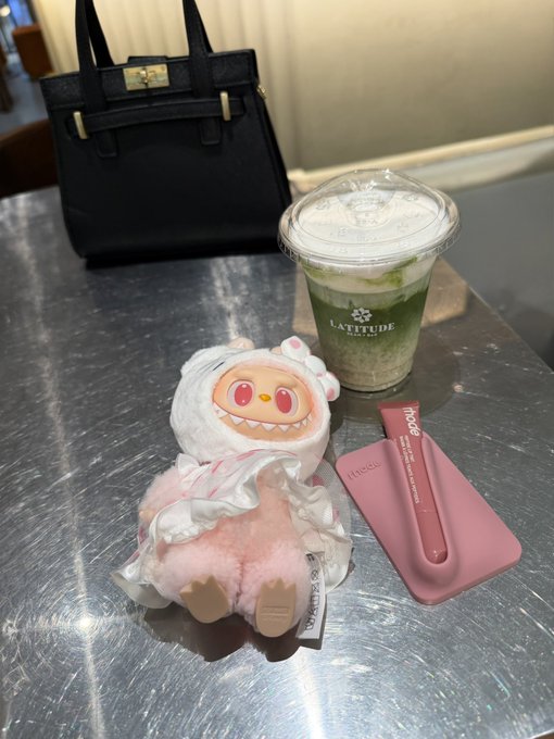A photo of a small plush doll in a white fluffy hooded outfit sitting on a metal table next to a cold green beverage from 'Latitude' and a pink 'rhode' lip case, with a black Hermès-style handbag in the background.
