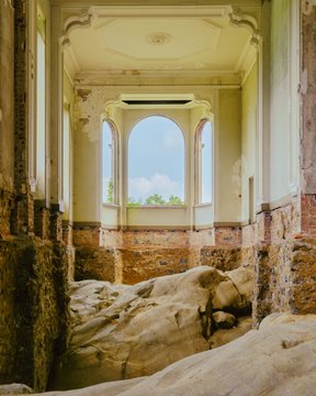 An interior view of an abandoned, decaying classical building featuring ornate pale walls, an arched window framing blue sky, and a floor buried under a thick layer of sand or debris.