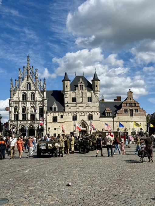 A historic European town square featuring Gothic-style buildings with spires, a vintage military jeep, people walking, and various flags displayed under a partly cloudy blue sky.