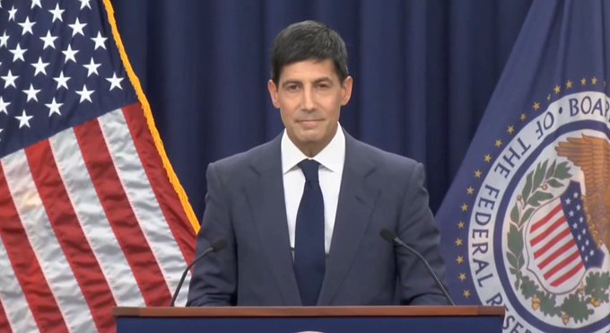 A man in a dark suit and tie stands at a podium, flanked by an American flag on the left and a seal of "THE BOARD OF THE FEDERAL RESERVE" on the right, suggesting a Federal Reserve official addressing markets ahead of the closely watched U.S. Producer Price Index (PPI) data release.