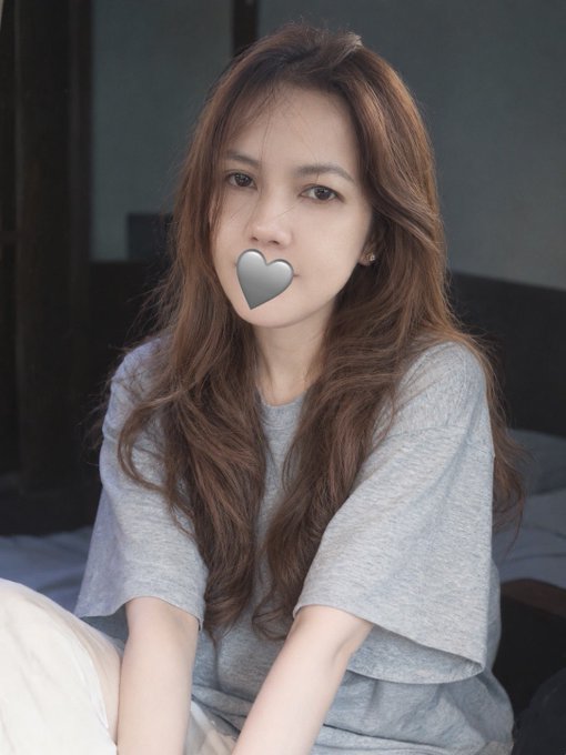 A young woman with long brown hair sits wearing a gray t-shirt, with a heart emoji covering her mouth.