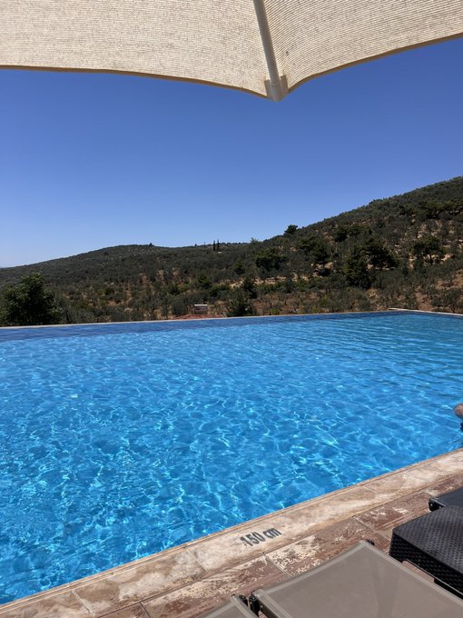 A view of an infinity pool with bright blue water overlooking a hillside of trees under a clear sky, with a "1.60 cm" depth marker visible at the edge and a sunshade above.