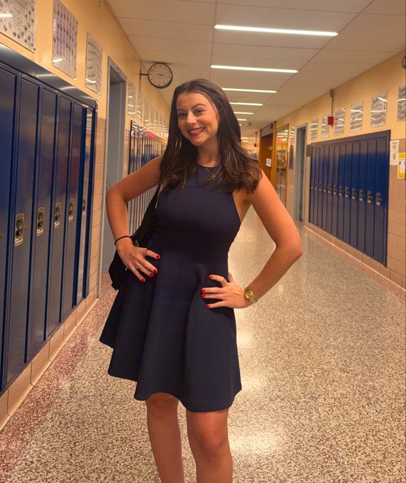 The image features a young woman wearing a blue dress, standing in an empty hallway with her hands at her sides. She has red nail polish on her nails and appears to be posing for the photo. The background of the image shows several lockers lining up against the wall, suggesting that she is in a school setting or a similar environment.