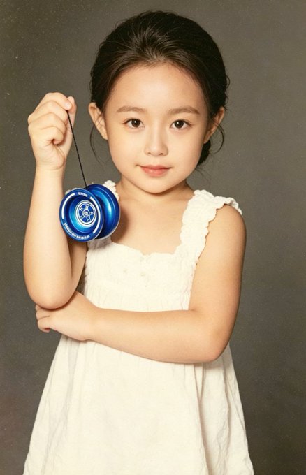 The image features a young girl holding a blue toy, which appears to be a toy wind-up toy or a yo-yo. She is dressed in a white dress and is posing for the picture with her hands on her hips. The background of the photo is black, making it stand out against the white dress. There are no other people visible in the image, so it seems like she is alone at the moment.
