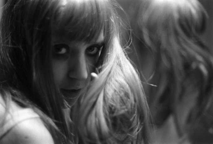 A moody black and white photograph captures a young woman with bangs gazing intensely at the camera, her reflection visible in a mirror behind her.