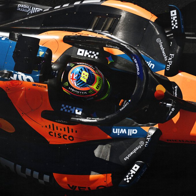 A McLaren Formula 1 driver, identified as Piastri by the name on the cockpit, sits in the car wearing a helmet featuring colorful designs and Monster Energy branding. The car is adorned with sponsor logos including OKX, Cisco, Dell, and Freshworks, capturing the moment as the driver prepares to ascend to the pinnacle of motorsport—fitting for a caption declaring "On top of the world."