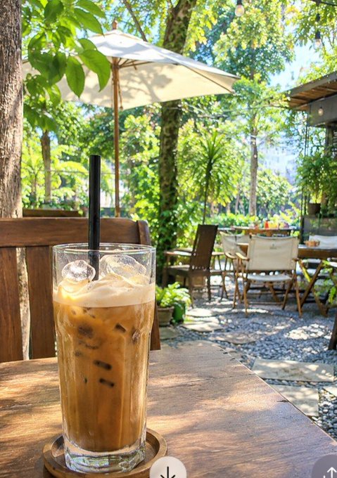 An iced coffee drink with a black straw sits on a wooden table at an outdoor cafe, with an umbrella, chairs, and lush green trees in the background.