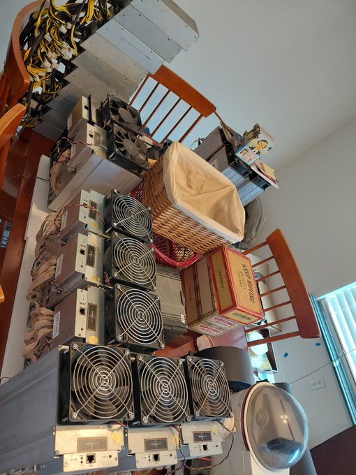 A home mining setup with multiple ASIC miners featuring large cooling fans stacked on furniture alongside a basket and cardboard box, with cables visible above.