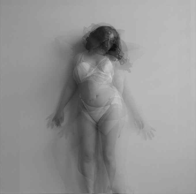 An artistic black-and-white photograph of a figure in white lingerie and a veil, with ghostly motion blur creating multiple silhouettes.