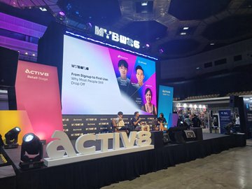 A view of a large convention hall featuring a stage with massive 'ACTIV8' lettering, with a presentation slide displayed on the screen behind the stage.