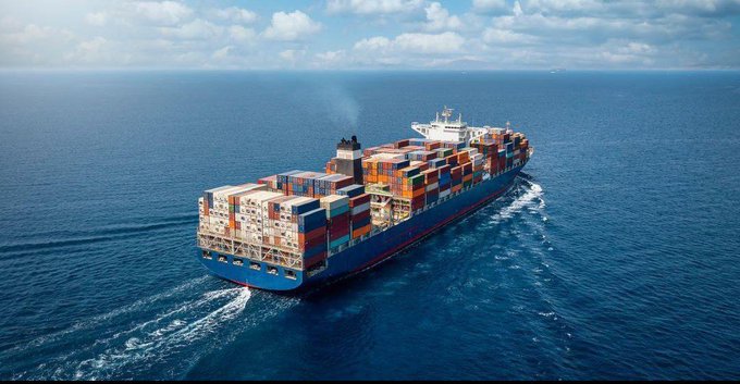 An aerial view of a large blue container ship loaded with multicolored shipping containers sailing across the open ocean.