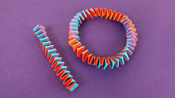 A colorful 3D paper origami bracelet in a zigzag pattern of red, orange, pink, and teal rests against a purple background, with a separate strip of the folded design placed beside it. The piece appears to be a creative craft representation possibly symbolizing ten years and connecting to the Bitcoin (₿) cryptocurrency anniversary celebration mentioned in the post.