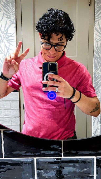 A mirror selfie of a man with curly dark hair and glasses, wearing a pink polo shirt, making a peace sign while holding a smartphone with a ring light attachment.