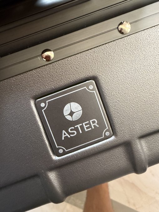 A close-up of a black merchandise case displaying the "ASTER" logo with its distinctive emblem, likely one of the limited-edition items mentioned for conference attendees in Indonesia.