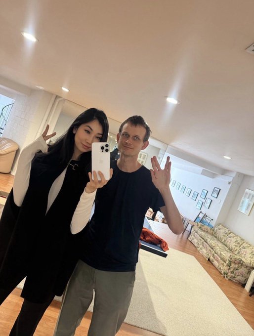 The image features a man and woman standing together, both holding up their phones to take a selfie. The man has his arms outstretched while the woman also has her hands raised for the picture. They are posing in front of a couch that is placed against one wall with a chair nearby. The background includes a rug on the floor and several pictures hanging on the wall above them, creating an atmosphere of a cozy living space.