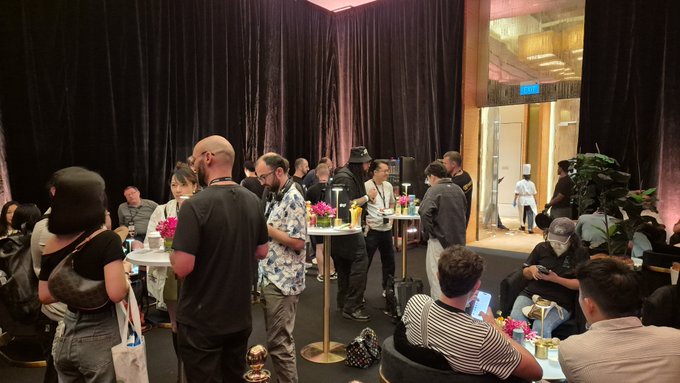 Guests mingle with drinks and conversation at high-top tables in a dimly lit venue with dark curtains, embodying the caption's sentiment that the room itself set the stage while the attendees brought the energy to life. A blue exit sign glows in the background as people socialize, some seated on couches and others standing with cocktails in hand.