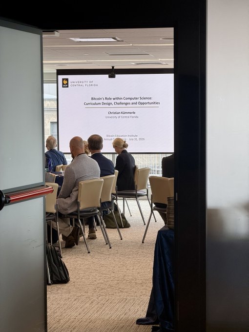 A photo taken through a doorway showing an audience seated in chairs facing a projection screen at the University of Central Florida, displaying a presentation titled 'Bitcoin's Role within Computer Science: Curriculum Design, Challenges and Opportunities' by Christian Kammel.