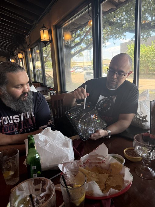 Two bearded men sit at a restaurant table by a window; one holds an audio mixer, while the other wears a Houston Football t-shirt, with chips, drinks, and a green bottle between them.