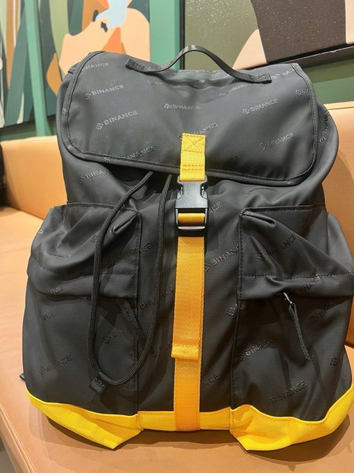 A black and yellow backpack branded with repeated 'BINANCE' logos sits on a tan chair.