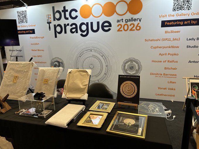 The image shows an art gallery display featuring various pieces of artwork, including paintings and sculptures. The table displays these items for sale or exhibition purposes. A sign reading "BTCPrague" can be seen behind the display, indicating that this event is taking place in Prague.