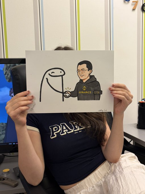 A woman wearing a navy 'PAUL' t-shirt holds up a framed cartoon illustration of a stick figure shaking hands with a man wearing a 'BINANCE' jacket and glasses.
