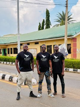 Three men wearing black 'RAVEN' t-shirts pose together on a street in front of a yellow building with a green roof.