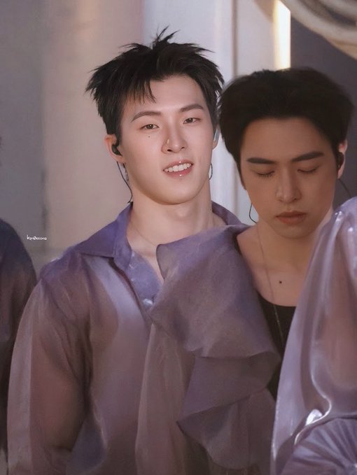 A photo of two young men wearing sheer lavender/purple tops, with the one in front smiling and wearing an earpiece, while the one behind has his eyes closed.