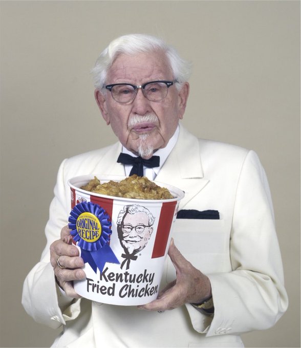 An elderly man with white hair and glasses dressed in a white suit and bow tie, resembling Colonel Sanders, holds a large bucket of Kentucky Fried Chicken branded 'Original Recipe'.