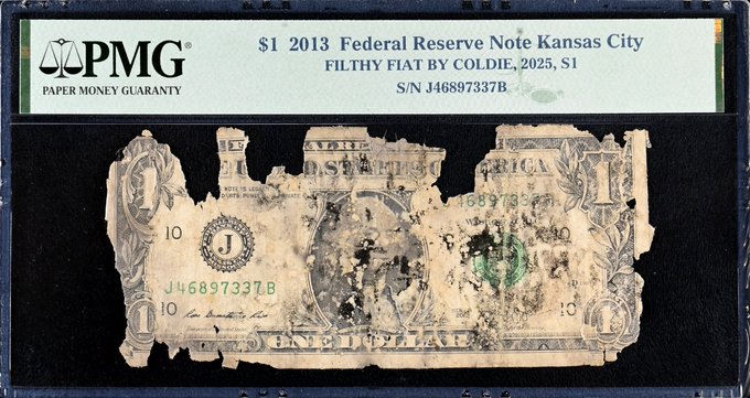 A PMG-graded encapsulated 2013 $1 Federal Reserve Note from Kansas City, labeled "Filthy Fiat by Coldie, 2025," showing a heavily damaged/collaged one dollar bill with serial number J46897337B.