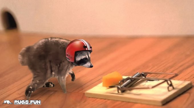 A kitten in a red football helmet approaches a mousetrap with cheese bait on a wooden floor, with a 'RUGS.FUN' watermark in the corner.