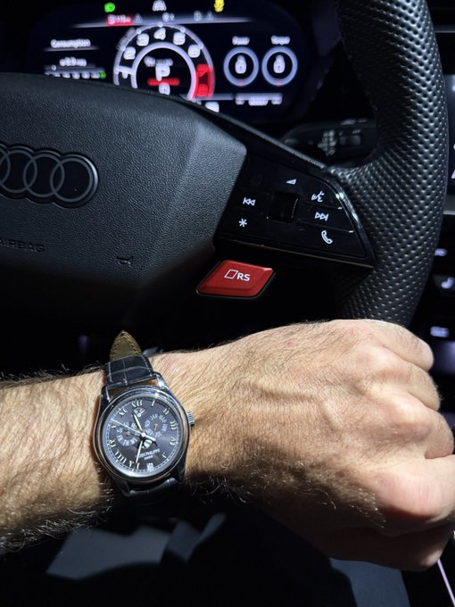 A man's wrist sporting a classic luxury chronograph watch rests on the steering wheel of an Audi, with the dashboard display and a red "RS" badge visible in the background—fitting for the "Watchkingliquid" caption by showcasing a high-end timepiece paired with a performance car.