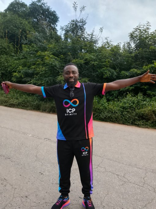 A man stands outdoors on a road with his arms outstretched, wearing a black polo shirt and pants featuring 'ICP DFINITY' logos in colorful neon hues, with lush green trees in the background.