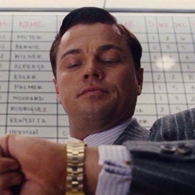 Leonardo DiCaprio as Jordan Belfort in *The Wolf of Wall Street* smirks smugly while checking his gold Rolex, with a whiteboard behind him listing names like "Wiener," "Alperna," and "Koplowitz"—a scene that perfectly captures the post's ironic joke about impatient investors imagining themselves living lavishly once their holdings pay off.