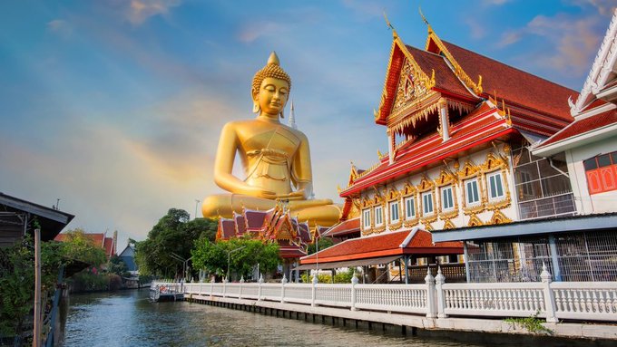 A golden Buddha statue sits beside an ornate Thai temple with red-tiled roofs beside a canal, with a rainbow arcing through the cloudy sky. The image accompanies the claim that Thailand has announced 0% capital gains tax on Bitcoin and crypto, illustrating the country's location tied to the tax policy news.