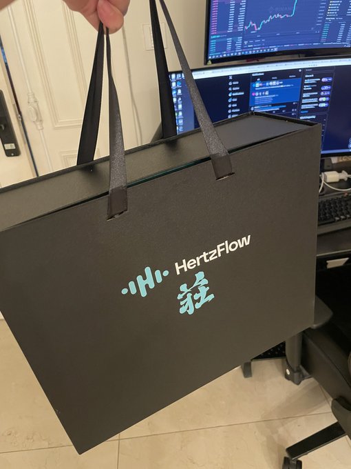 A hand holds up a black shopping bag featuring the "HertzFlow" logo in light blue lettering alongside Chinese characters, presented as a gift received from @Hertzflow_xyz. In the background, dual monitors display trading charts and market data on what appears to be a trader's desk setup.
