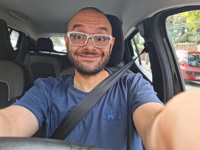 A selfie of a smiling bald man with glasses and a beard, wearing a blue t-shirt and seatbelt, sitting in the driver's seat of a car.