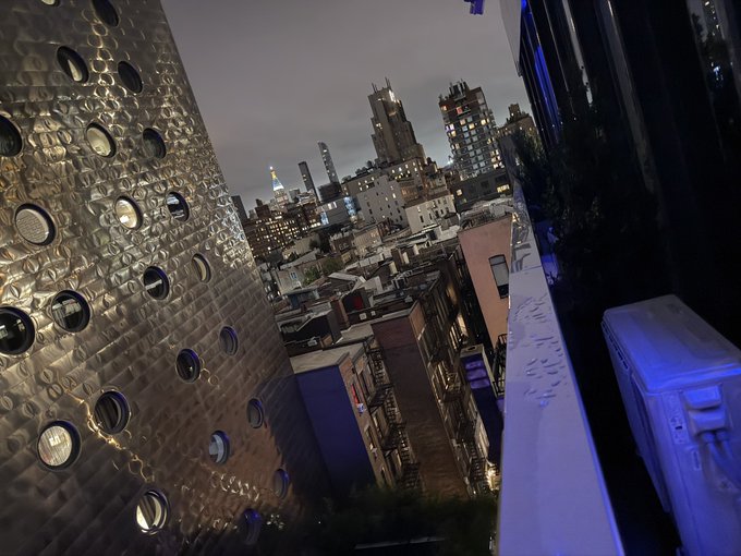 A nighttime cityscape viewed from an elevated outdoor space, showing a metallic perforated wall with circular cutouts on the left and city buildings including what appears to be the Empire State Building in the distance.