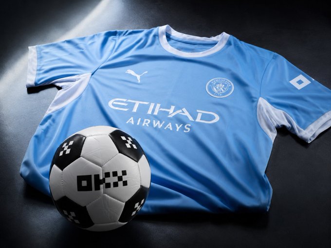 A light blue Manchester City football jersey sponsored by Etihad Airways with a Puma logo lies next to a black and white soccer ball featuring a pixelated pattern on a dark surface.