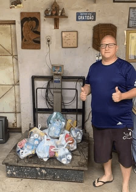 A man in a blue t-shirt and dark shorts gives two thumbs up while standing next to a large industrial scale loaded with multiple bags of plastic bottles for recycling.