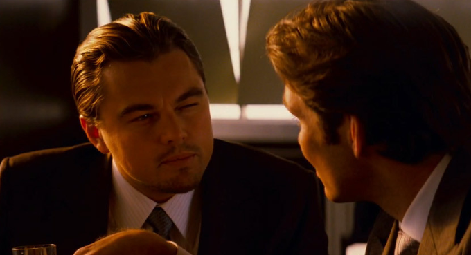 Leonardo DiCaprio in a dark suit looks intently at another man in what appears to be a scene from a movie, likely "Inception."
